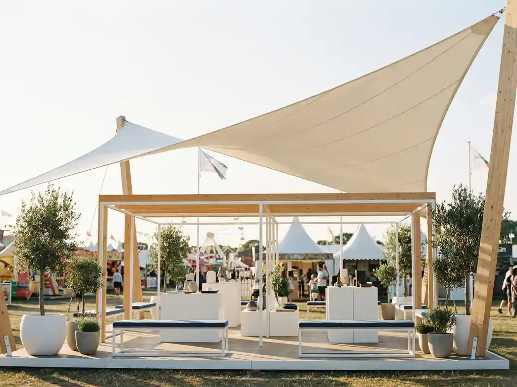 Moderner Messestand im Freien mit weißem Sonnensegel-Baldachin, hellem Ausstellungsmobiliar und Naturholzakzenten unter blauem Himmel.